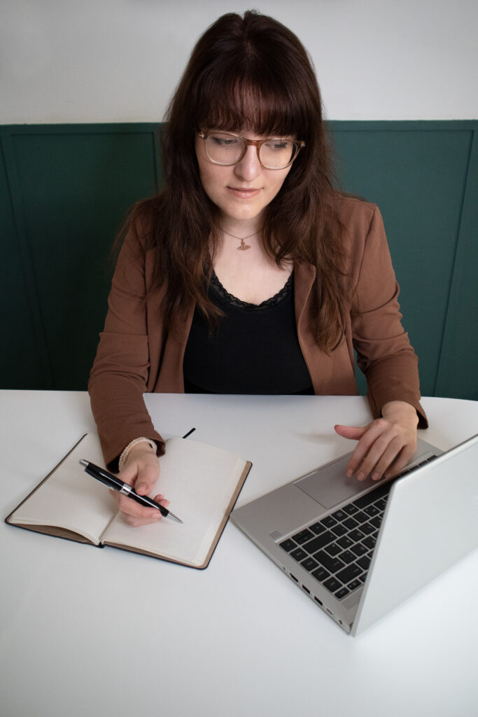 Die freie Texterin sitzt an einem Tisch. In der rechten Hand hält sie einen Kugelschreiber über ein offenes Notizbuch, mit der linken Hand fährt sie über das Touchpad eines Laptops.