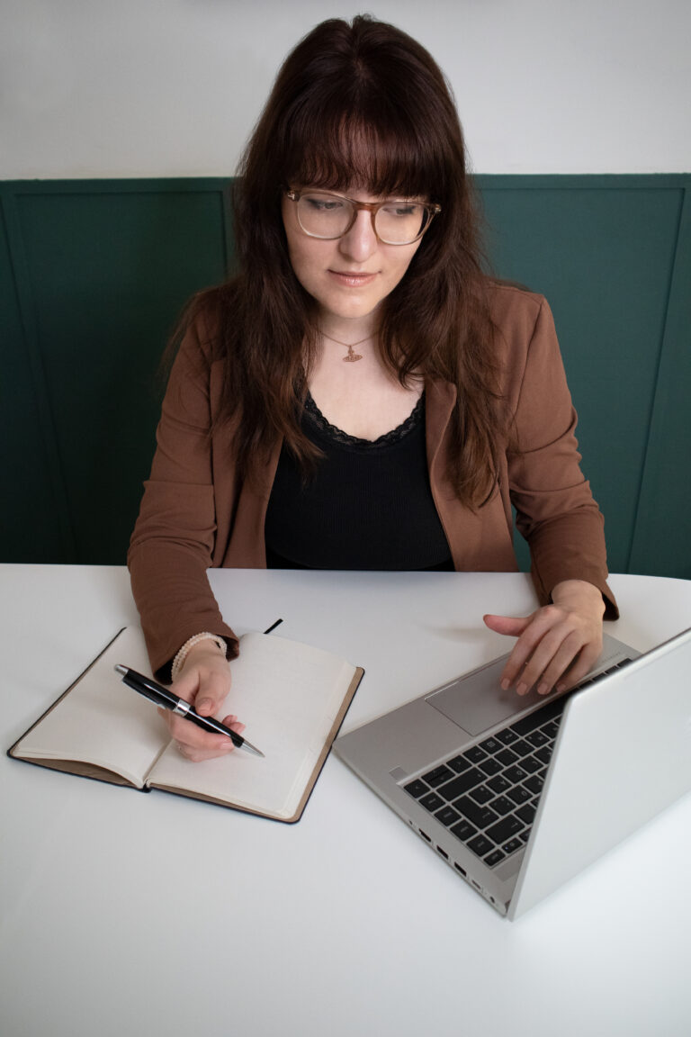 Die freie Texterin, Sarah Kreuzer, sitzt am Tisch und schaut in ein Laptop. Mit einer Hand hält sie einem Kugelschreiben über einem offenen Notizblock.