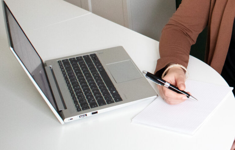 Bildausschnitt, auf dem ein Laptop und ein Notizzettel zu sehen sind. Eine Hand, die einen Kugelschreiber hält, befindet sich über dem Blatt.