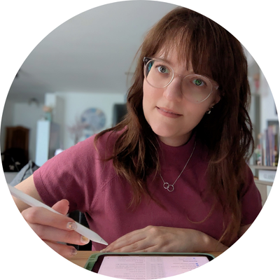 Die freie Texterin, Sarah Kreuzer, sitzt mit ihrem iPad am Tisch und hält den iPen in der Hand. Sie schaut lächelnd in die Kamera.