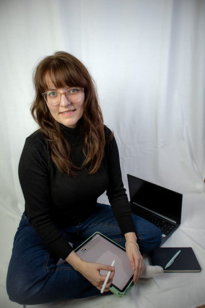 Die freie Texterin, Sarah Kreuzer, sitzt im Schneidersitz auf dem Boden und lächelt in die Kamera. Sie schreibt mit einem iPen auf ihrem iPad, neben ihr liegen noch ein Notizbuch und ihr Laptop.