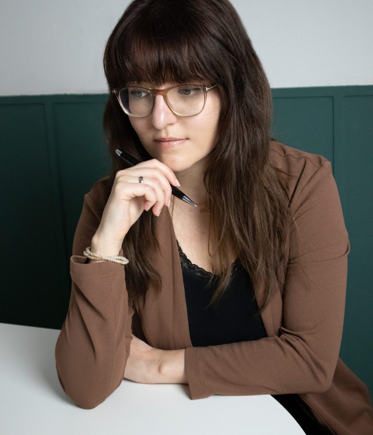 Die freie Texterin, Sarah Kreuzer, sitzt an einem Tisch. Sie hat die Hand an ihrem Kinn, darin befindet sich ein Kugelschreiber. Außerdem blickt sie sanft lächelnd zur Seite.